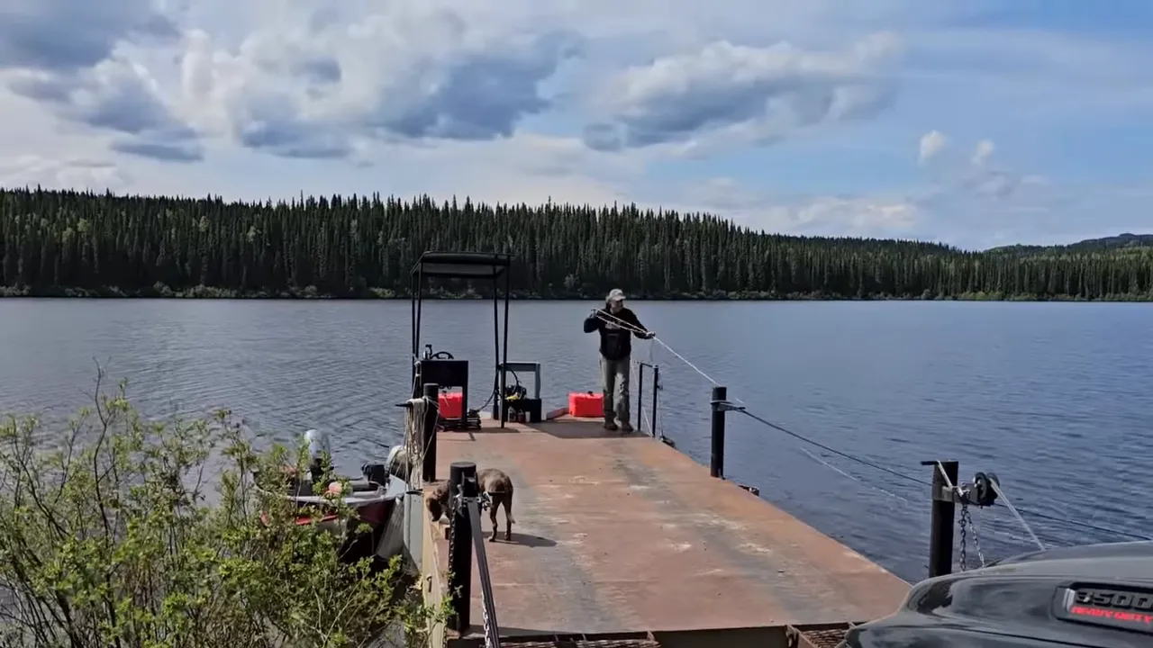 Person handling a bow line on the barge with a dog on the ramp and the lake and treeline in the background