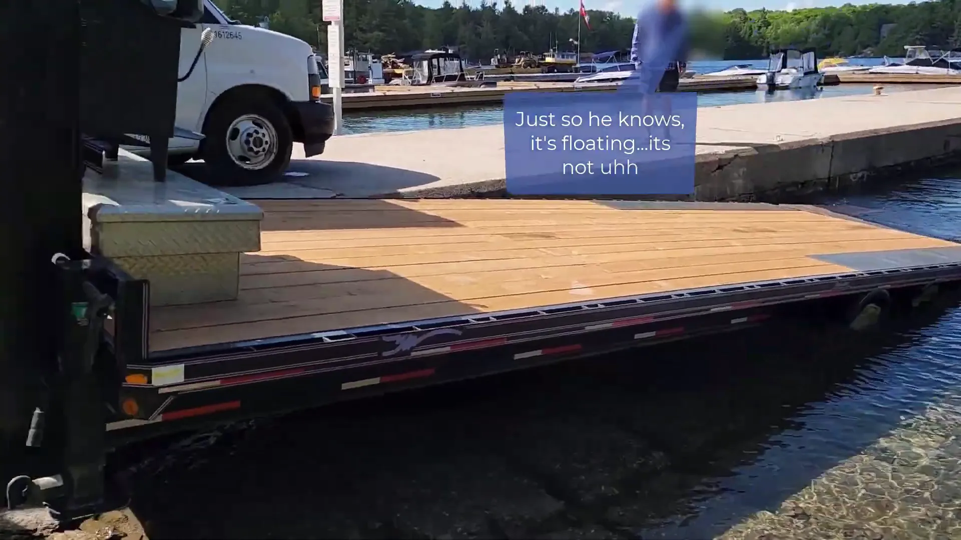 A large trailer is backing up into a lake beside a dock