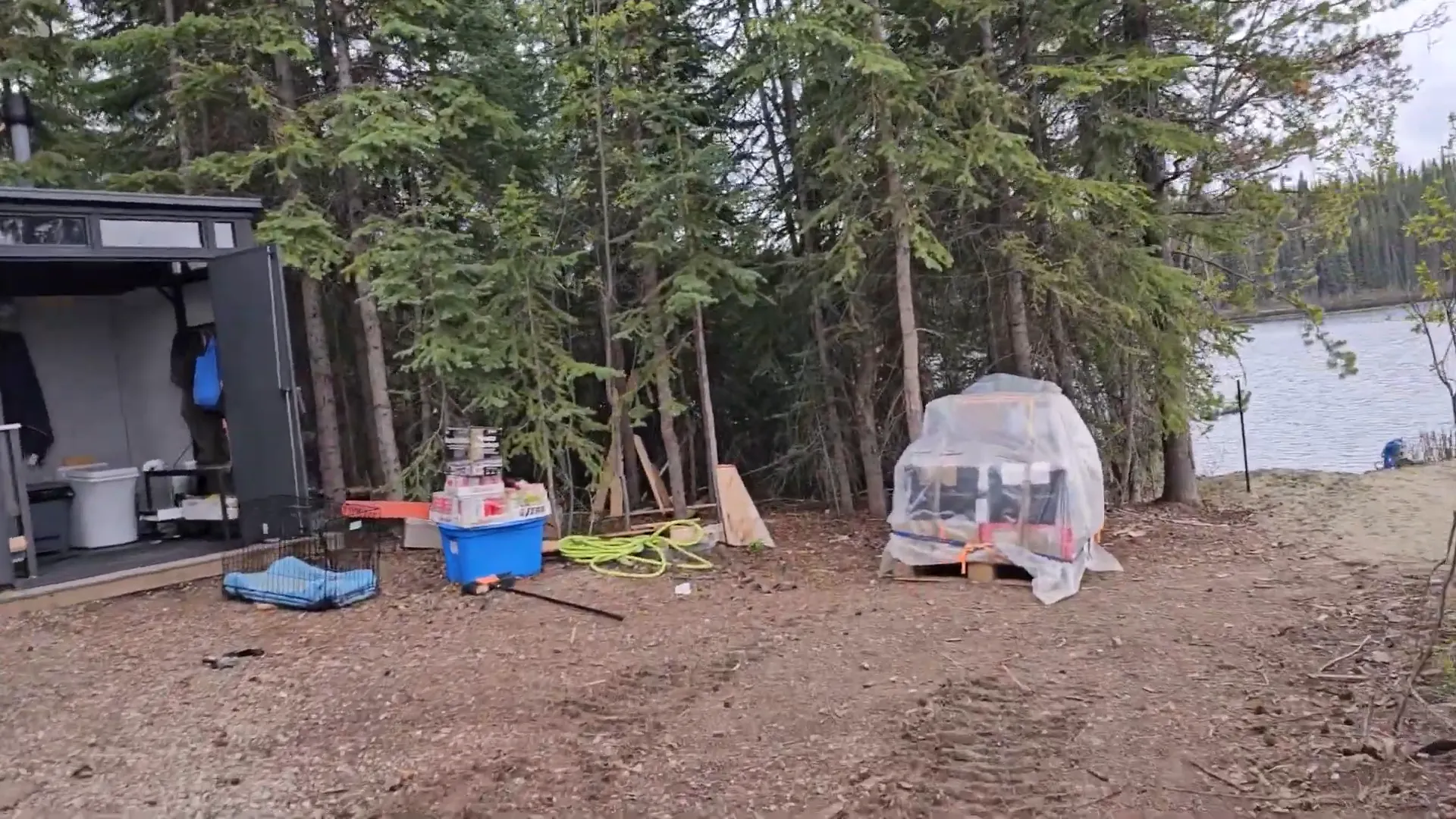 Off-Grid Island Winter Damage: What’s Still Standing? | S3 Ep2 2 A campsite with a pallet of supplies, a bathroom shed next to a lake