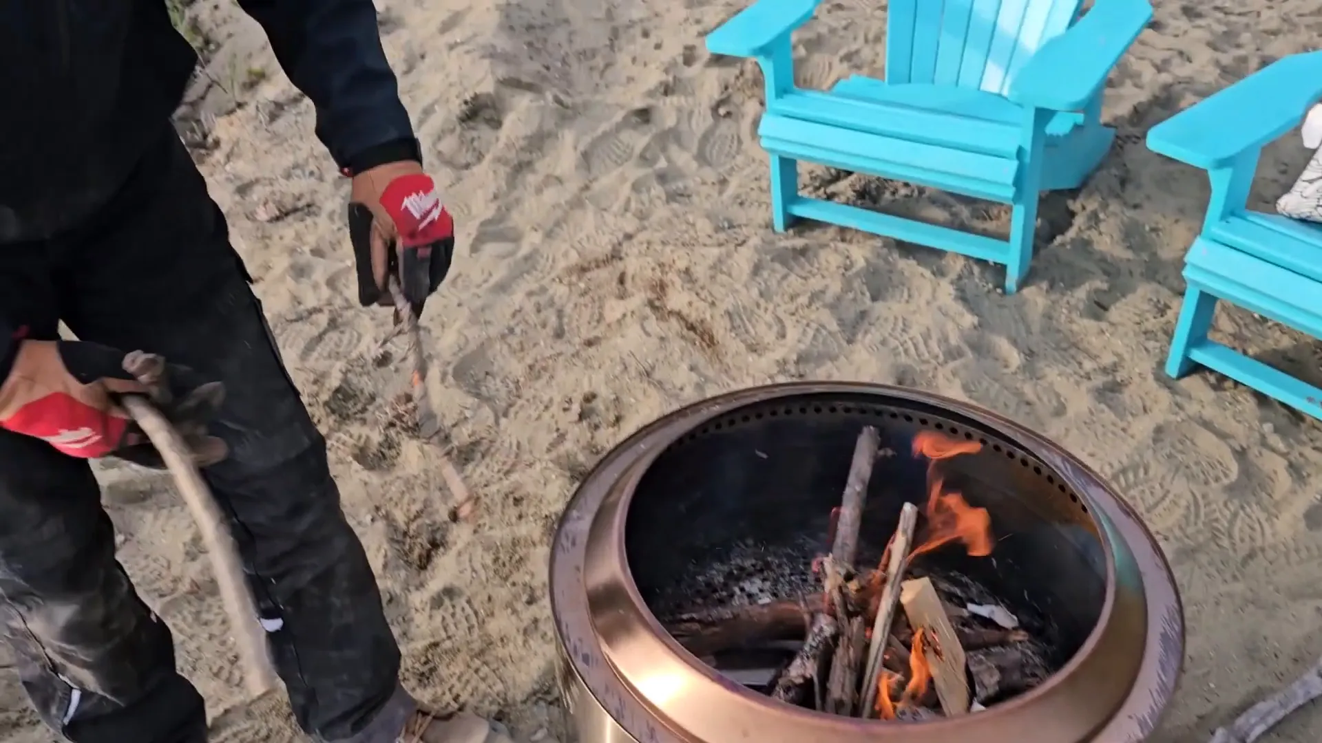 A man wearing gloves tends to a fire in a stainless steel pit