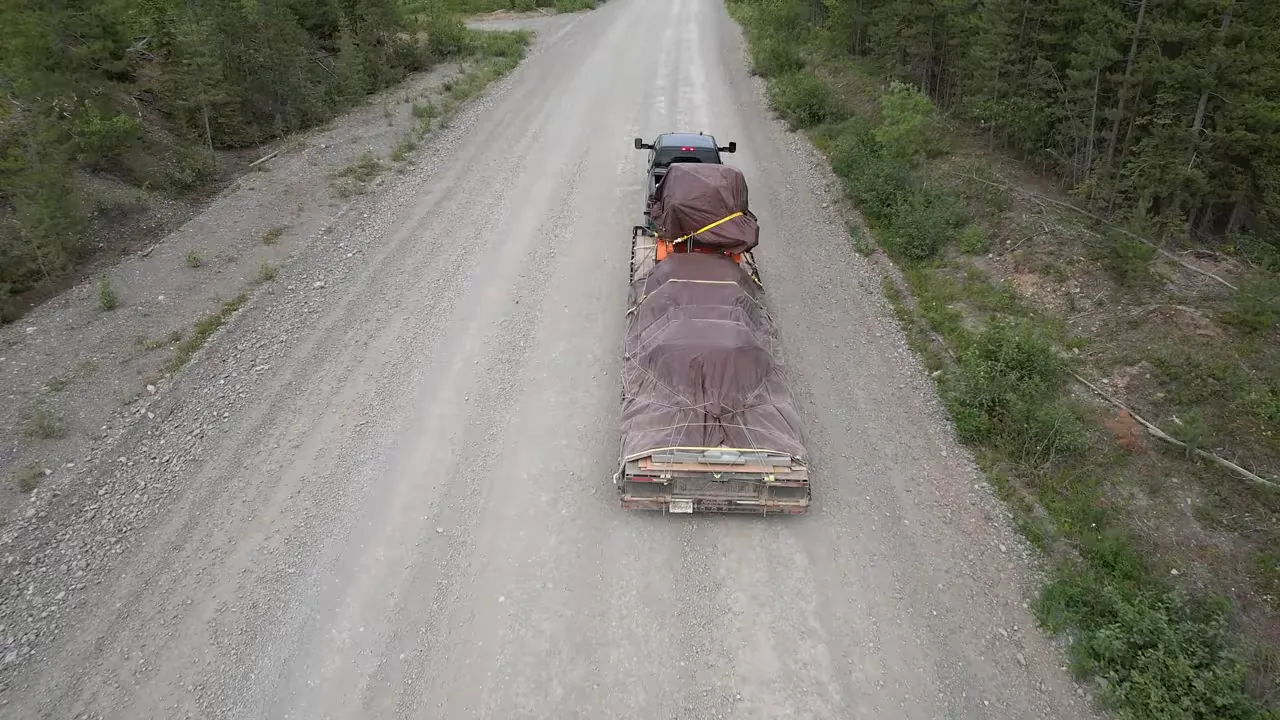 One Slip and Our New Sawmill Falls Into the Water! - S4 Ep11 3 Top-down aerial of a pickup towing a covered sawmill on a washboard gravel road with visible straps