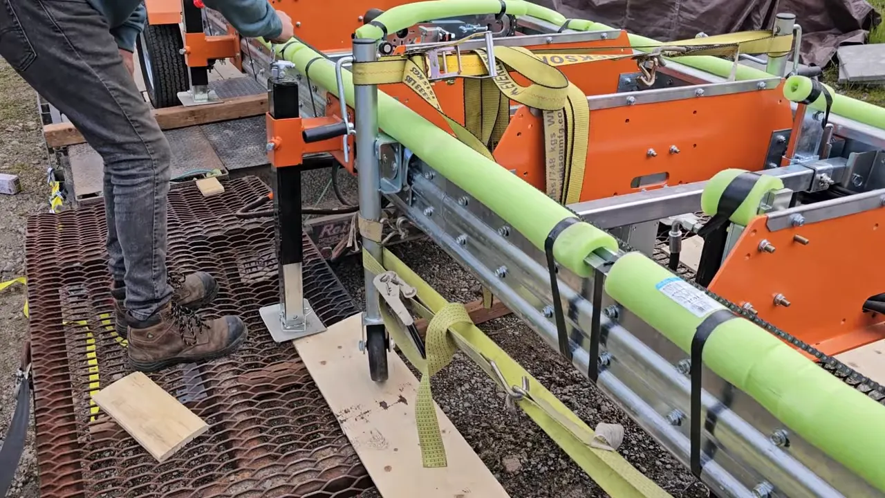 One Slip and Our New Sawmill Falls Into the Water! - S4 Ep11 8 Close-up of a portable sawmill frame with yellow ratchet straps, pool-noodle padding, a screw jack, small caster wheel and wooden blocks on a grated ramp