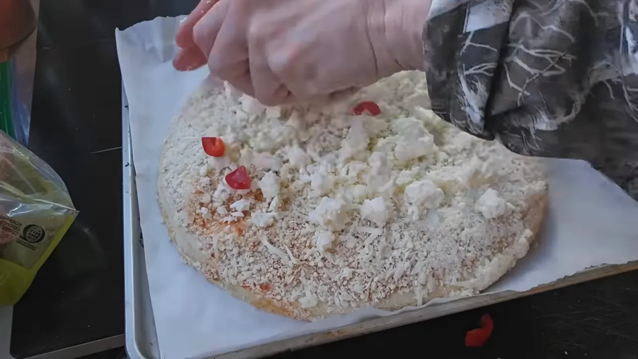 Hands sprinkling crumbled feta over a pizza with small pieces of red bell pepper visible