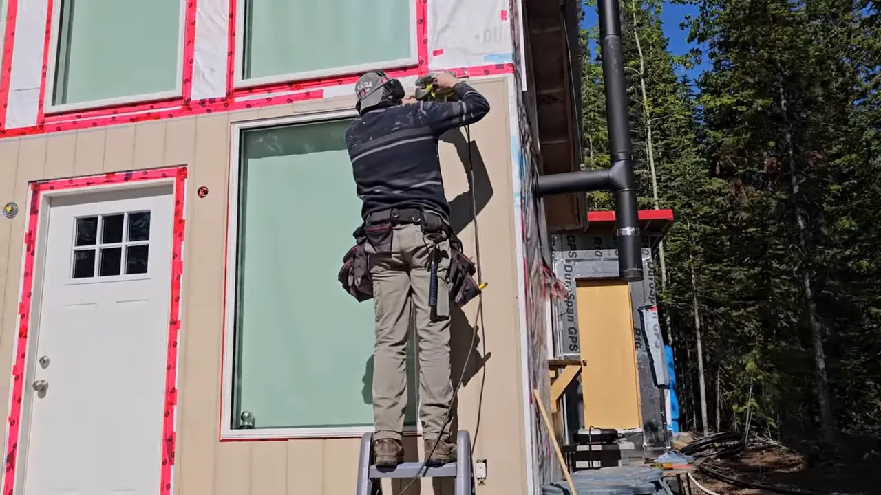 Off Grid Life Canada: Building a Tiny Cabin on Our Remote Island – Biggest Transformation Yet! - S4 Ep8 4 Worker standing on a ladder holding a circular saw overhead to cut siding around a large window on a tiny cabin.