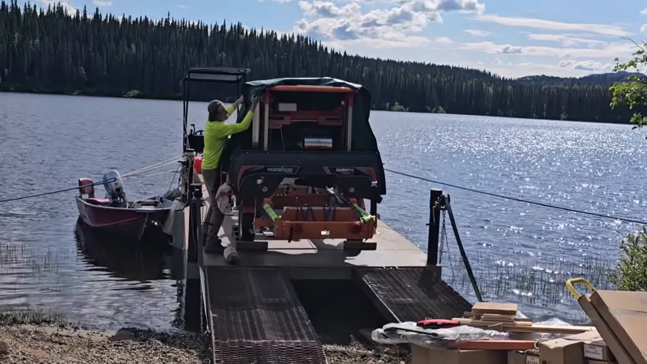 Front-on view of norwood sawmill on a barge with person adjusting a tarp; pool noodles visible on the mill's frame and ramps leading to shore.