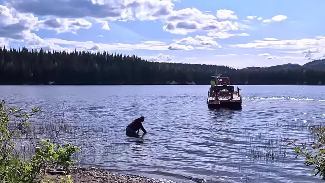 Person crouched in shallow lake water adjusting a pump line with a barge in the background