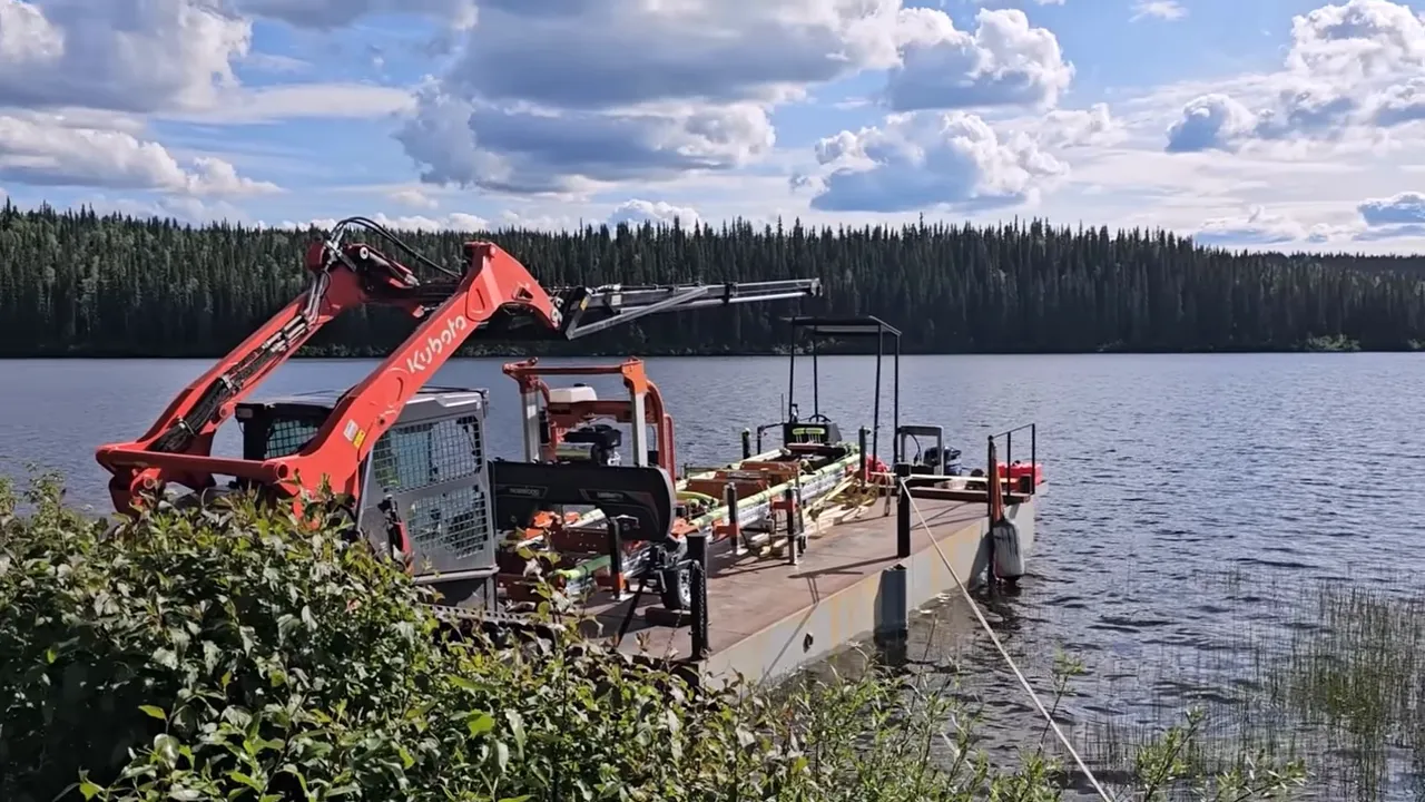 Wide shot of a barge with a kubota boom lift extended toward the shore against a forested lake backdrop