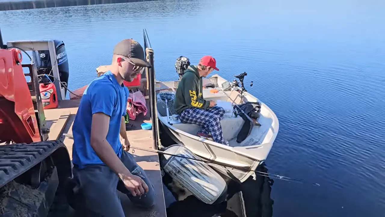 Two men on a skiff at a barge dock with supplies on a remote lake