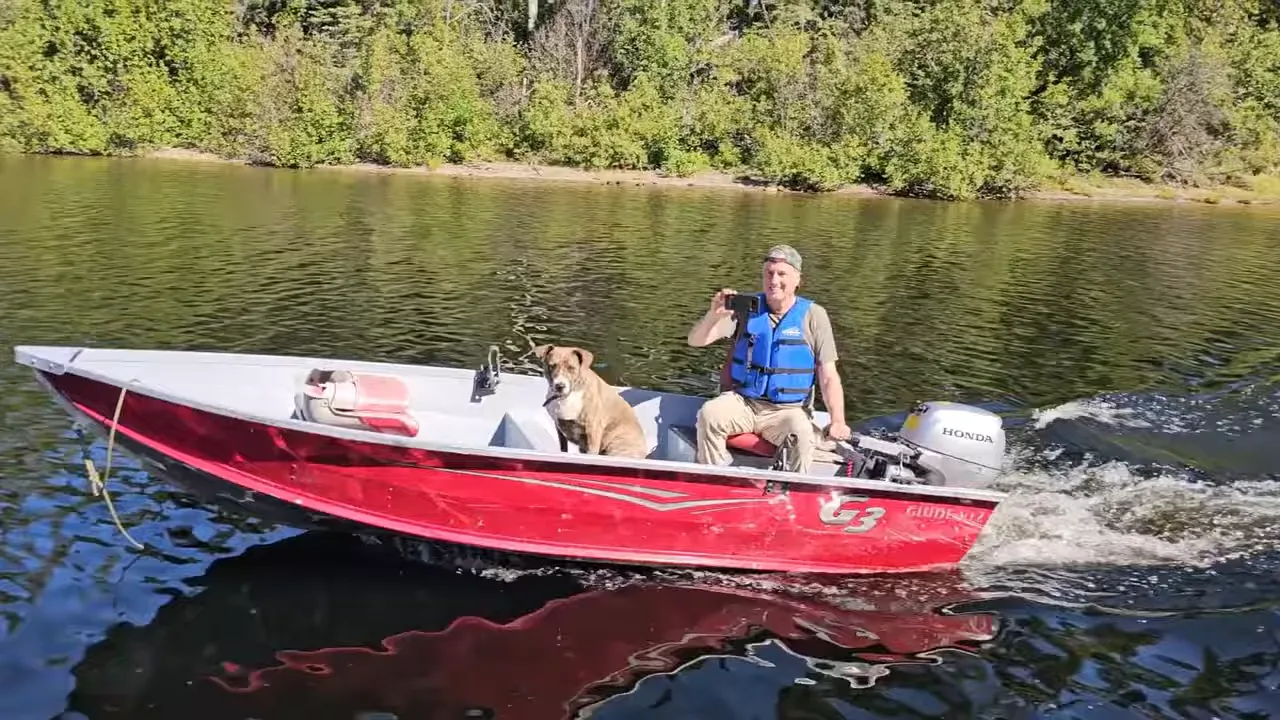 Red skiff with a person at the helm and a dog riding on a calm remote lake