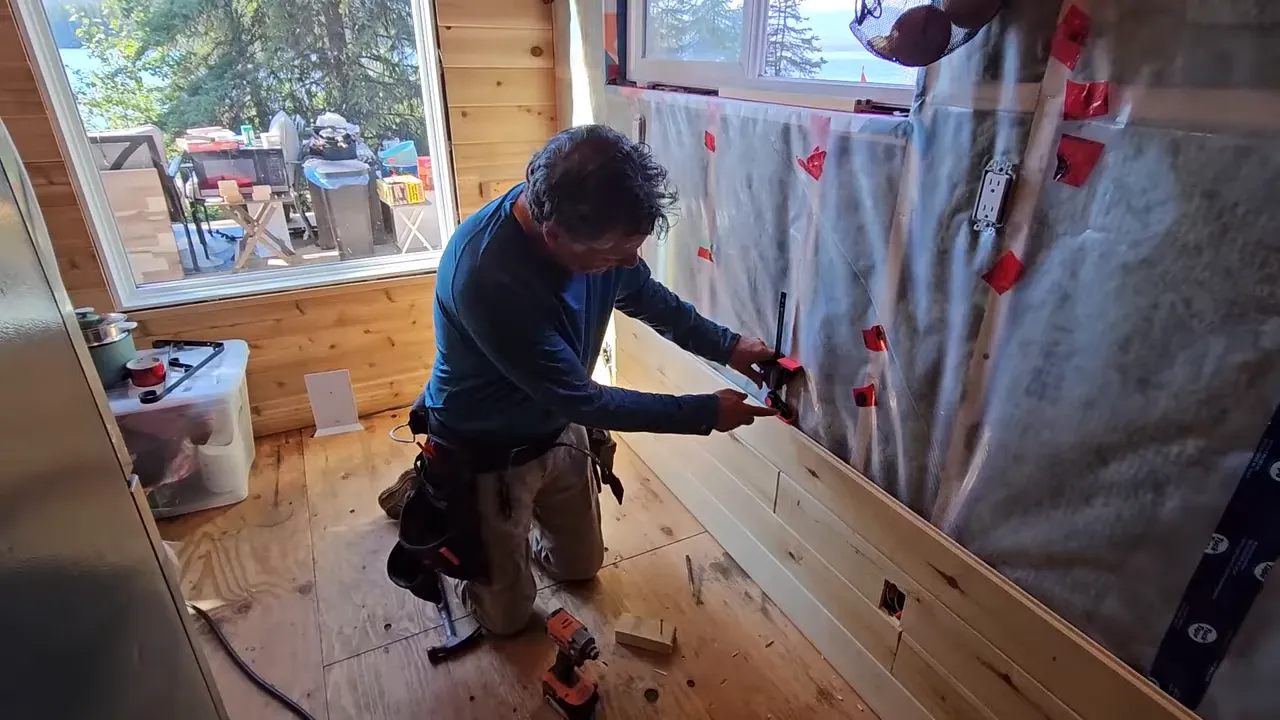Builder securing an off-grid cabin interior wall while tools sit on the floor and vapor barrier is visible