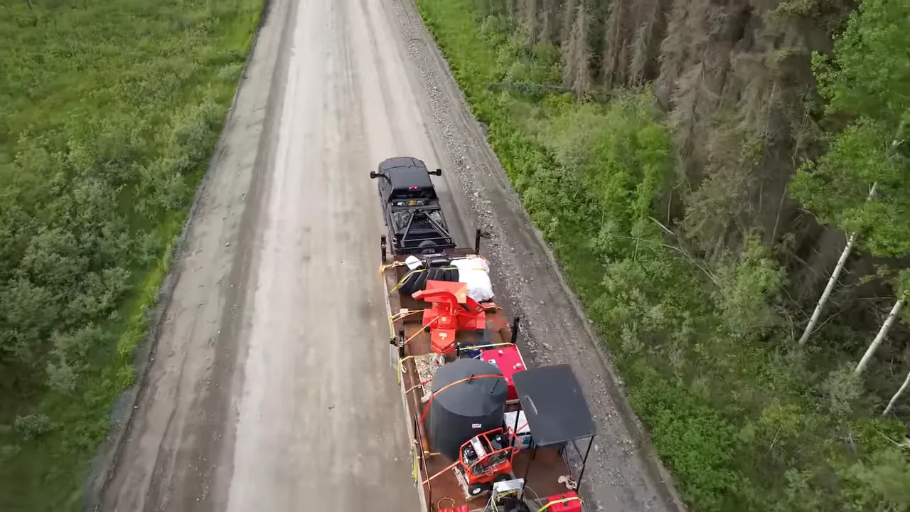 Aerial shot of a truck towing a heavily loaded trailer along a gravel road through dense forest, showing equipment strapped down.