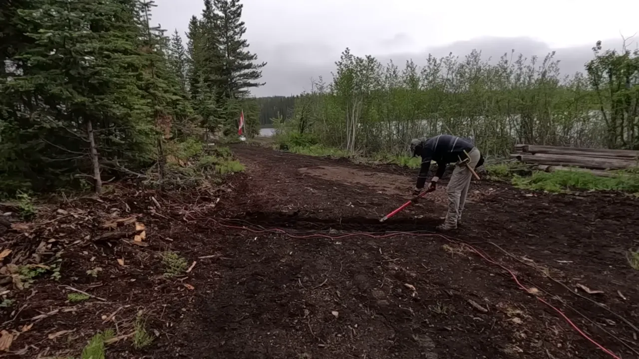 Wide view of a cleared dirt access route with a person working to position red and black electrical cables across a shallow trench; lake and trees visible in the background.
