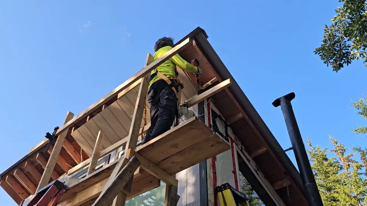 Worker on a tied-in harness standing on wooden scaffolding installing soffit trim under a cabin eave