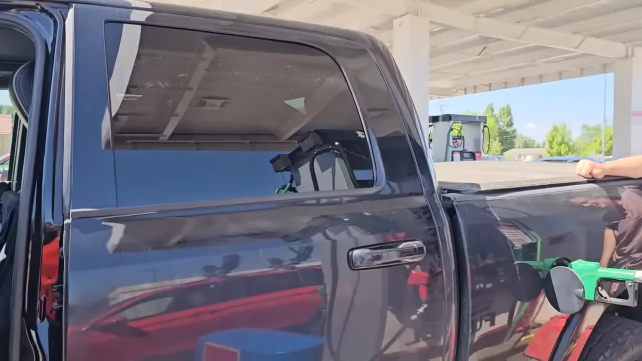 Black pickup truck being refueled at a gas station with a green fuel nozzle inserted under the station canopy