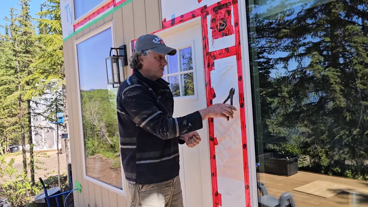 Off Grid Life Canada: Building a Tiny Cabin on Our Remote Island – Biggest Transformation Yet! - S4 Ep8 5 Worker holding nippers beside taped window and door trim with tree reflections and deck visible