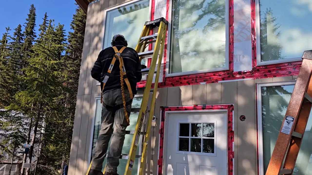 Off Grid Life Canada: Building a Tiny Cabin on Our Remote Island – Biggest Transformation Yet! - S4 Ep8 12 Worker climbing a ladder and fitting siding on the front of a tiny cabin with taped window reveals and a second ladder nearby