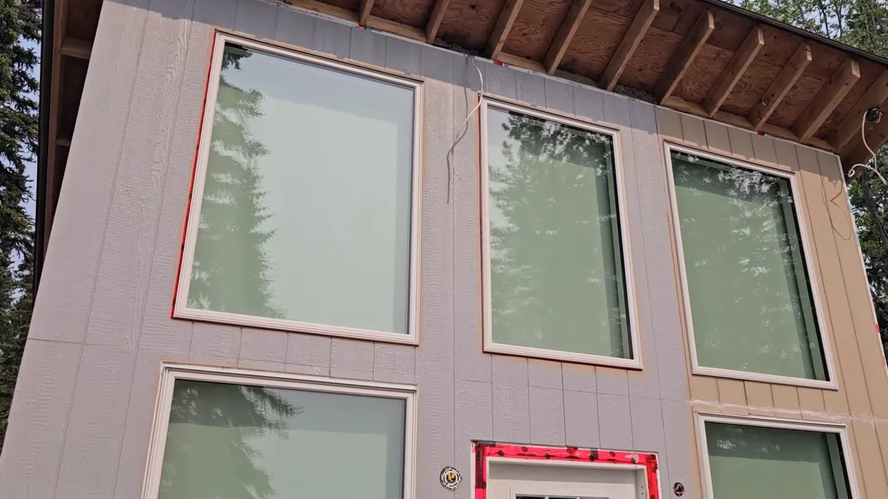 Off Grid Life Canada: Building a Tiny Cabin on Our Remote Island – Biggest Transformation Yet! - S4 Ep8 10 Front of tiny cabin with muted gray painted siding, three large windows with tree reflections and red flashing tape around the door