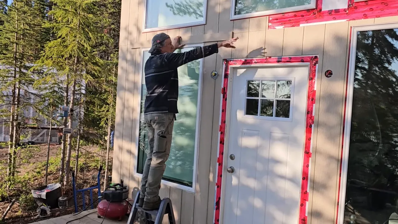 Off Grid Life Canada: Building a Tiny Cabin on Our Remote Island – Biggest Transformation Yet! - S4 Ep8 8 Person aligning and pointing at siding trim above a window and door with flashing tape ready for caulk